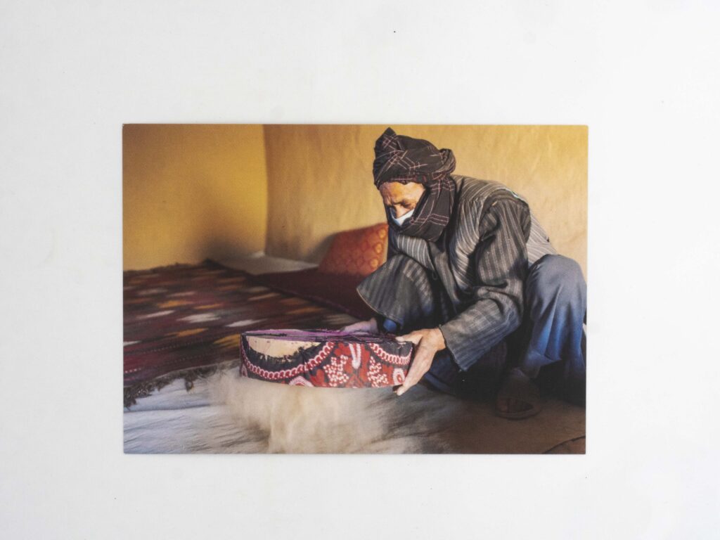 Cannabis postcard from Afghanistan: An Afghan man in traditional attire sieving dried cannabis plants to extract hashish in an outdoor setting, with cannabis materials spread on a tarp in the background.